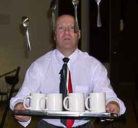 bald white man with red white & blue tie behind magic floating spoons with a tray of coffee cups